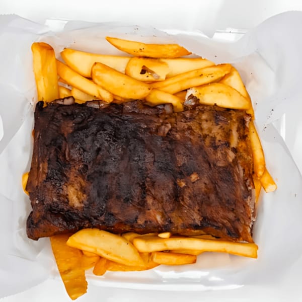 A rack of dark, smoky BBQ ribs with a side of golden french fries in a white container.